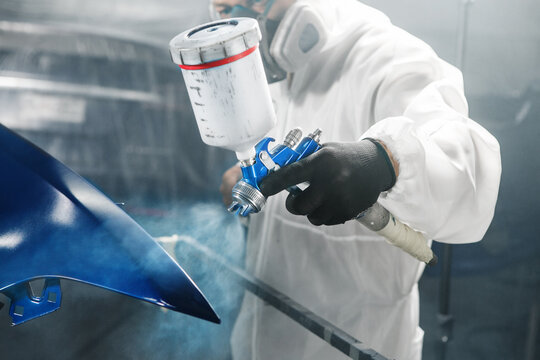 Male automobile repairman painter in protective workwear and respirator painting part of car body in paint chamber