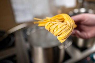 A chef's hand holds freshly made pasta above a pot of boiling water. The kitchen setting features stainless steel cookware, emphasizing a culinary preparation scene.