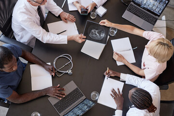 Top view of woman passing CT image of patients skull to chief physician discussing X ray examination results with colleagues, while sitting at table with laptops during meeting at medical office