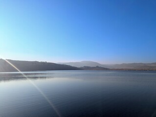 A serene view of a mountain lake captured on a sunny day, with distant peaks rising majestically along the horizon. The lake's surface mirrors the vivid blue of the sky, creating a harmonious blend 