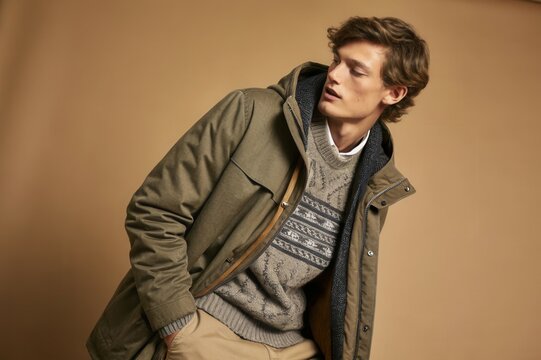 A young man poses confidently in a cozy, stylish outfit featuring a knitted sweater and a jacket, showcasing contemporary winter fashion against a warm, neutral backdrop during a photoshoot.