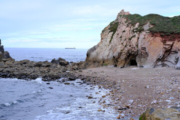 A rocky shoreline with a boat in the distance. The water is calm and the sky is overcast