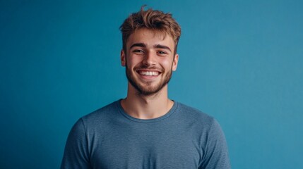 Fototapeta premium A cheerful young man stands against a solid blue backdrop, wearing a casual t-shirt. He appears friendly and approachable, embodying positivity in a real estate marketing context.