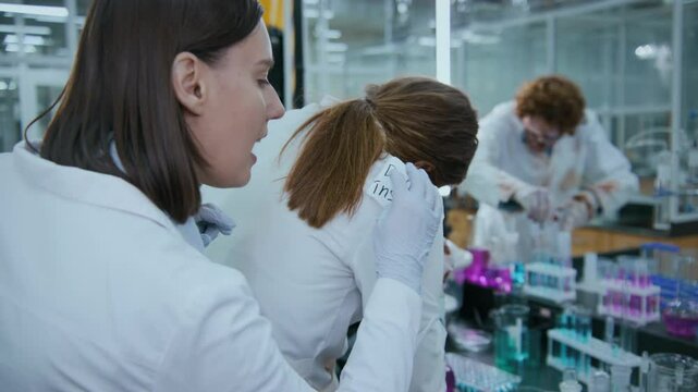 Medium shot of cheerful human female Caucasian lab technician pressing finger to lips, then placing taunting sticker on back of zombie colleague, who is studying test tubes and abusing equipment