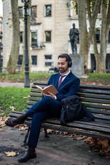 Obraz premium Businessman wearing headphones reading book while sitting on bench in park