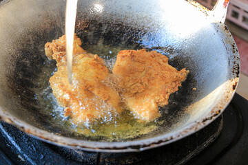 Two pieces of fried chicken are sizzling in hot oil inside a wok. Water is being poured over the chicken. Crispy Fried Chicken Cooking in a Wok.