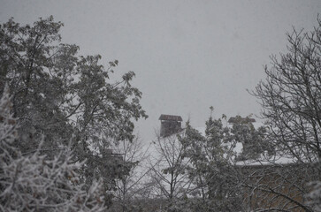 A snowy scene with a chimney on a house. The chimney is covered in snow and the trees are bare. Scene is peaceful and serene, with the snow covering the ground and the trees.