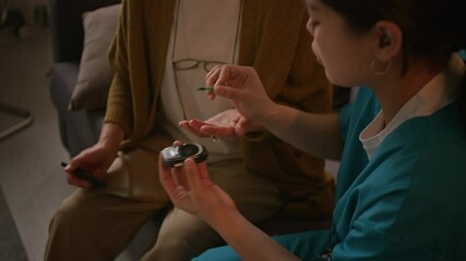 Young female nurse taking blood sample from unrecognizable elderly woman suffering from diabetes for checking sugar level with glucose meter, while sitting in living room during home care visit