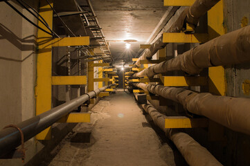 Underground concrete utility tunnel with pipes and wires lit by tungsten bulbs.. This tunnel gives...