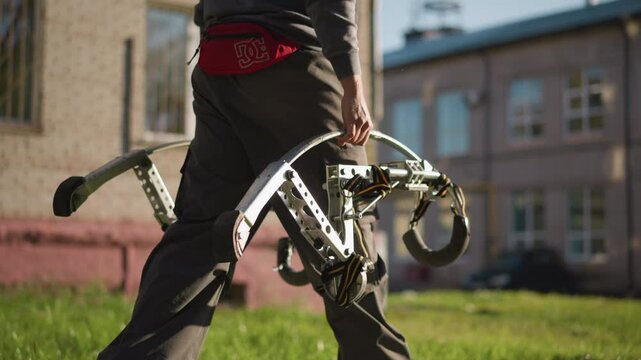 A Guy carrying large metallic spring stilts, wearing black pants, dark gray long-sleeved shirt, and red hip bag with visible white logo. Background is blurred with grassy area and distant building