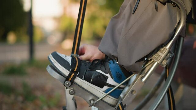 Close-up of person fastening shoes to spring stilts using orange and black straps, wearing gray trousers and long-sleeved shirt outdoors, blurred background, focus on adjustment process
