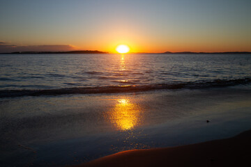 Fototapeta premium Golden sunset over calm ocean waves reflecting light, with a serene sandy shore in the foreground. Perfect for travel, nature, or peaceful scenery themes.