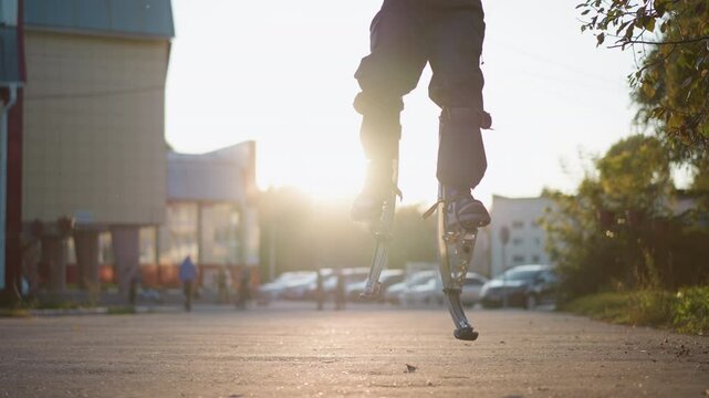 Person with short brown hair wearing gray hoodie and dark gray pants jumping on spring stilts on paved road in residential area surrounded by houses, showcasing unique motion and urban lifestyle