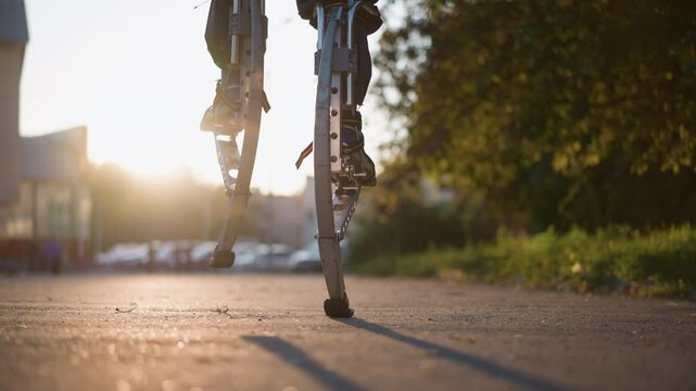 Close-up view of legs walking on spring stilts in suburban outdoor setting with dark pants and sneakers. Background features blurred trees, buildings, and a street. Low camera angle highlights