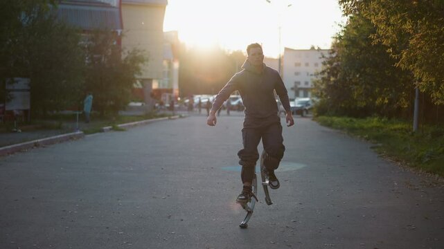 Person with short brown hair wearing gray hoodie and dark gray pants, using spring stilts on paved road in residential area. Captured from distance, with trees and buildings in background