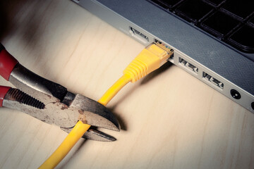 Sharp pliers severing yellow ethernet cable connected to laptop, dramatic angle. Stark composition symbolizing digital disconnection, censorship and network interruption.

