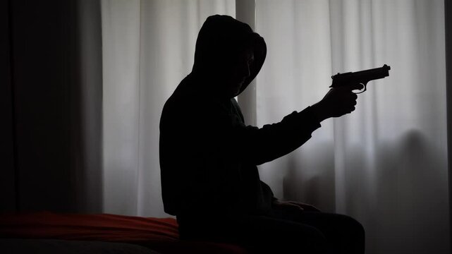Hooded man terrorist learns to use a revolver gun weapon at home, thief thug prepares a burglary robbery in apartment. social dangers connected to the increase in gun ownership in the USA America