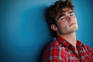 Fototapeta premium A young man leans against a blue wall, looking relaxed and casual