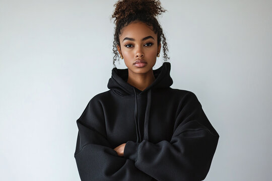 An african american female model wearing a black oversized designer hoodie, standing against a clean white backdrop.