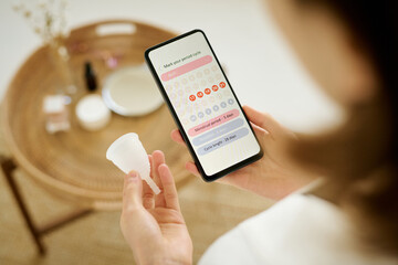 High angle shot of unrecognizable female hands holding white menstrual cup and smartphone with period calendar