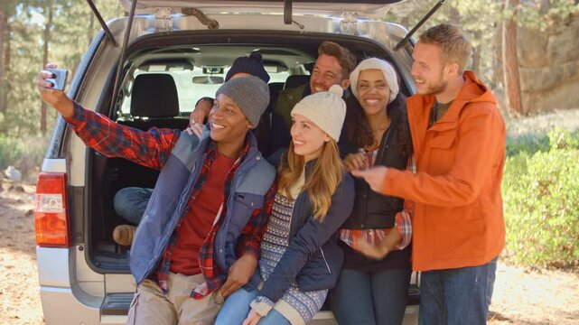 Group of young friends outdoors sitting in open tailgate of car posing for selfie on mobile phone before walk through autumn or fall countryside - shot in real time