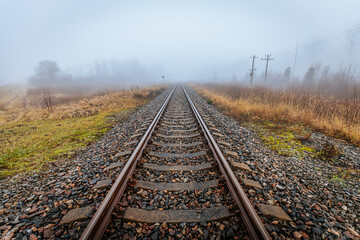 Obraz premium Straight railroad tracks surrounded by foggy fields and grass, leading into the horizon. The mist and muted colors evoke solitude and serenity.