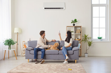 Young couple, happy family with dog on sofa switching on air conditioner, blowing warm, cold air...