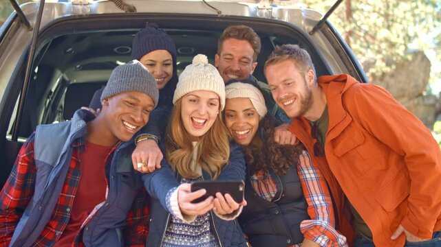 Group of young friends outdoors sitting in open tailgate of car posing for selfie on mobile phone before walk through autumn or fall countryside - shot in real time