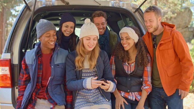 Group of young friends outdoors sitting in open tailgate of car posing for selfie on mobile phone before walk through autumn or fall countryside - shot in slow motion