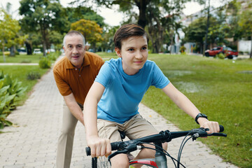 Obraz premium Cheerful boy riding a bike while his granddad pushing him from behind to teach him cycling