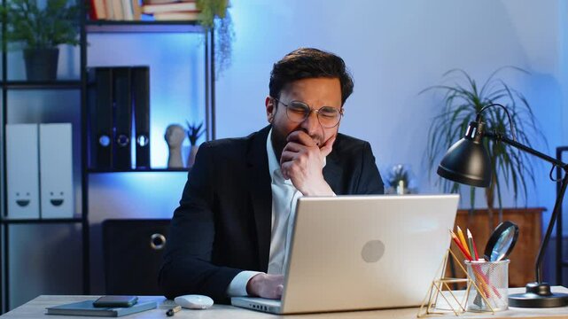 Bored sleepy Indian businessman worker working on laptop computer, yawning, leaning on hand falling asleep at office. Exhausted tired freelancer workaholic young man. Employment, occupation, workless