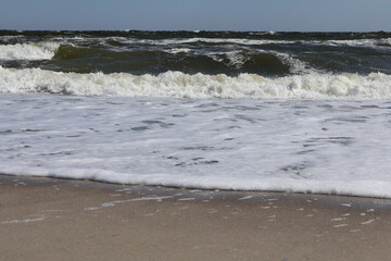 A dynamic coastal scene with rolling waves and white foamy water gently touching the sandy shore under a clear blue sky