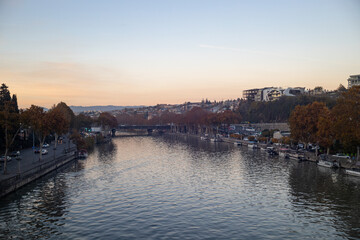 Obraz premium Serene autumn twilight envelops Tbilisi as golden leaves float by the river, painting a peaceful urban landscape.
