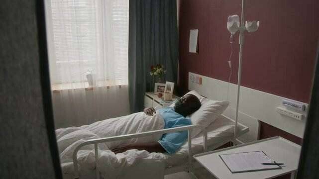 High angle shot of elderly black man in blue medical gown, with breathing mask over his face, lying under white blanket on single bed next to IV stand in hospital ward