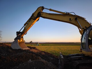 A crawler excavator stands at the edge of a field in the evening light. The view stretches to the horizon