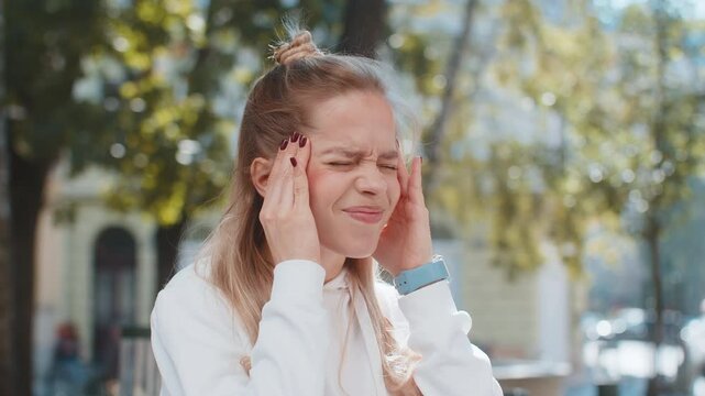Displeased exhausted Caucasian woman rubbing temples to cure headache problem suffering from tension migraine stress. Young female girl teenager tourist grimacing, pain standing in park city street.