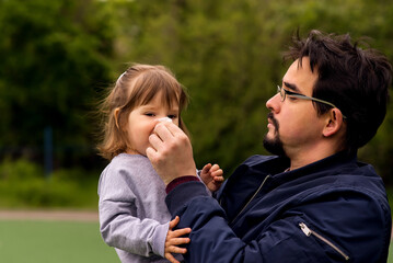 Fototapeta premium Middle-aged father in glasses wiping nose to his little child with white paper handkerchief outdoors in spring or autumn. Green leaves in background. Parental care and health protection concept