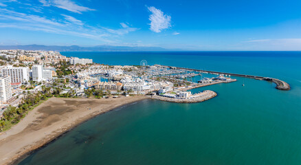 Fototapeta premium The image captures Benalmadena's marina with boats, sandy beaches, and white Mediterranean buildings. A Ferris wheel stands near the vibrant blue sea.