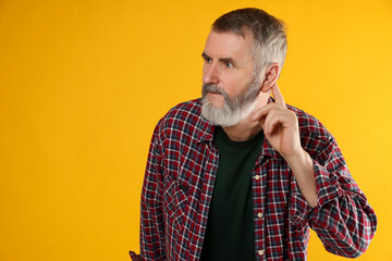Senior man showing hand to ear gesture on orange background, space for text