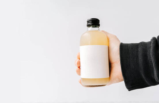 Woman hand holding small transparent glass bottle with black cap and white label, filled kombucha of yellow
