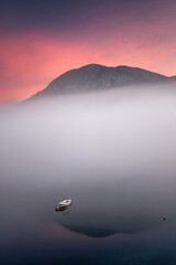 Serene morning scene with a small boat on calm water, a misty mountain, and a beautiful pink sky