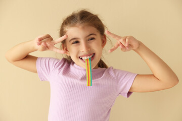 Happy girl eating tasty rainbow sour belt and showing peace signs on beige background