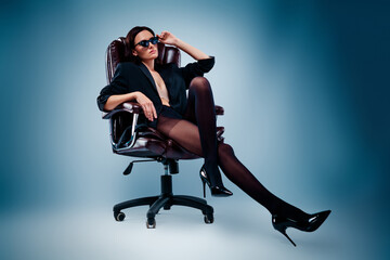 Confident young woman in a black jacket poses stylishly on a chair with a blue gradient background.
