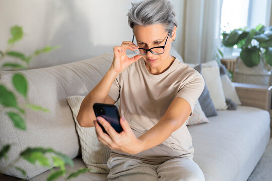 A middle-aged woman sits on the couch and lifts her glasses trying to read a message on the phone.
