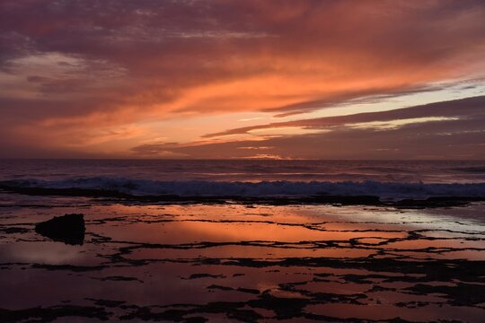 Golden sunset at Playa de Mangueta Beach, Spain, reflecting fiery torn up cloud structures in soft orange and light blue on the still waters of circular rock pools