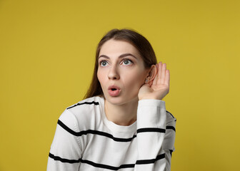 Woman showing hand to ear gesture on dark yellow background