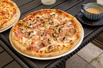 Delicious full fresh mushroom and ham pizza served on outdoor restaurant table next to a ceramic coffee cup, close-up food photography of Italian cuisine meal on a plate outdoors, outside, no people