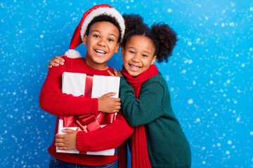 Fototapeta premium Photo of funny sweet dark skin kids dressed sweaters headwear scarf holding gift smiling hugging isolated blue color background