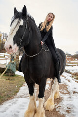 Obraz premium woman princess in black cloak sits astride black steed horse. Background winter nature.