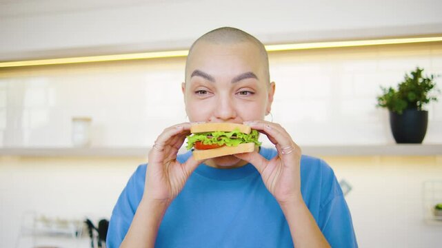 A bald woman, a cancer survivor, enjoys a hearty sandwich in her cozy kitchen on a sunny morning. A touching moment of strength and gratitude.